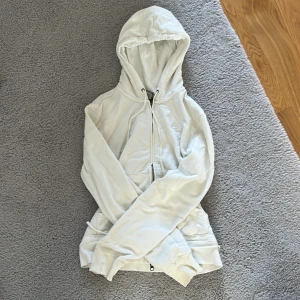 Vit hoodie från Nike - Säljer en stilren vit hoodie från Nike med dragkedja och broderad logga på bröstet. Den har en praktisk huva och fickor framtill. Den är i L men passar m också! Kan användas av xs och s men är lite större