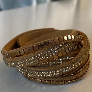 Brunt armband med glittriga detaljer!  - Snyggt brunt armband med flera rader av glittriga stenar och guldiga detaljer. Armbandet har en praktisk tryckknapp för enkel justering och passar perfekt för att ge en extra touch till din outfit. Jättefint armband  3 knappar för olika storlekar  Strasset/pärlorna är guldiga  Totala längden är 39 cm  Aldrig använt