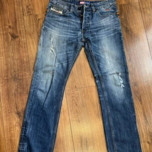 Blå jeans från Diesel - Fråga bara så löser jag fler bilder. Snygga blå jeans från Diesel med en klassisk femficksdesign och slitna detaljer. Jeansen har en knappgylf och en broderad logga på bakfickan. Perfekta för en avslappnad stil.