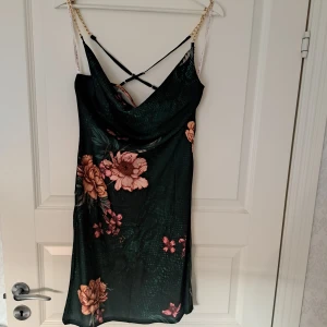Blommig klänning från Guess - Snygg blommig klänning från Guess i mörkgrön nyans med rosa blommor. Klänningen har tunna axelband med kedjedetaljer och en v-ringad design med korsade band i ryggen. Perfekt för en somrig look! Aldrig använd,  inköpt i GUESS butik i Florida