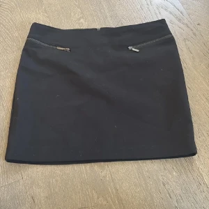 Svart kostymkjol lågmidjad från Wera Stockholm - Snygg svart lågmidjad kostym kortkjol från Wera Stockholm med dragkedjor framtill. Perfekt för en stilren look. Kjolen har en enkel design som gör den lätt att matcha med olika toppar.