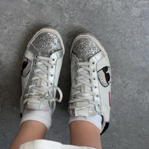 Vita sneakers från Karl Lagerfeld med glittriga detaljer - Snygga vita sneakers från Karl Lagerfeld med glittriga silverdetaljer på tån och hälen. Skorna har en unik design med ett tryck av Karl Lagerfelds ansikte på sidan och märkesnamnet i rosa. Perfekta för att ge din outfit en extra touch av glamour. Köptes på Vestiaire förra året i nyskick för 2000, nypris ca 3000. Pris kan diskuteras 💝