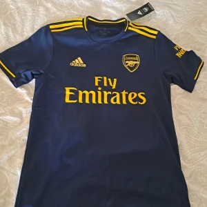  Arsenal fotbollströja  - Snygg mörkblå Arsenal fotbollströja från Adidas med gula detaljer och korta ärmar. Tröjan är så ny som de kan vara, Fick den som present från släktingar i USA men för liten tyvärr.