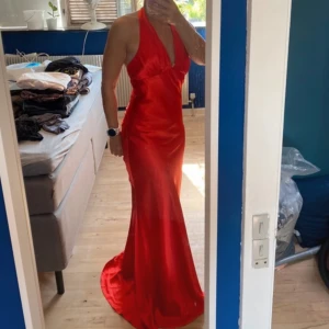 Röd långklänning i satin - Snygg röd långklänning i satin med halterneck och djup v-ringning. Perfekt för en elegant kväll. Klänningen har en smickrande passform och en vacker glans.