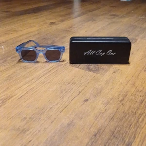 Blå solglasögon från All Cap One - Snygga blå solglasögon med transparent båge från All Cap One. Perfekta för soliga dagar. Kommer med ett svart fodral och putsduk för enkel förvaring och rengöring. I fint skick skriv om du har några funderingar!