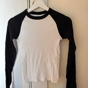 Vit och svart långärmad tröja från Brandy Melville (One size) - Säljer en stilren vit och svart långärmad tröja med ribbad struktur. Perfekt för en avslappnad look. Tröjan har en klassisk raglan-design med svarta ärmar och vit kropp.