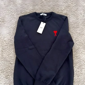 Svart tröja från Ami Paris - Snygg svart tröja från Ami med ett rött hjärta och bokstaven A broderat på bröstet. Tröjan har en klassisk rund hals och långa ärmar. Perfekt för en stilren look. Hör gärna av dig vid frågor eller funderingar! Mvh Melker