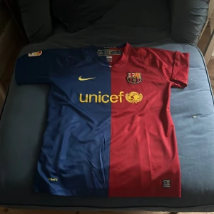 Blå och röd fotbollströja från Nike - Säljer en klassisk FC Barcelona fotbollströja från Nike i blått och rött med korta ärmar. Tröjan har klubbens emblem på bröstet och Unicef-loggan i gult. På baksidan finns namnet och numret 10 i gult och messi tröja, jsg har köpt den här tröjan när jsg och min pappa vart i Barca och kollad! 
