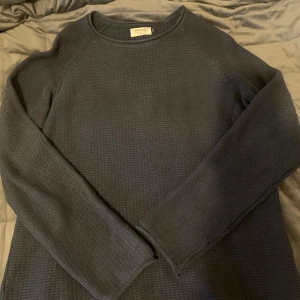 Mörkblå stickad tröja från Jack & Jones -  mörkblå stickad tröja från Jack & Jones. Tröjan har en klassisk rund halsringning och långa ärmar. Lite sliten vid halsen syns knappt 