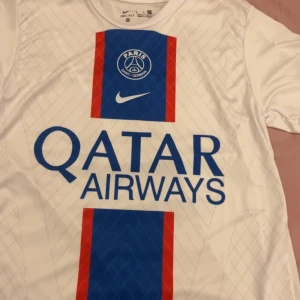 Vit PSG fotbollströja från Nike - Säljer en vit PSG fotbollströja från Nike med blå och röda detaljer. Tröjan har korta ärmar och är tillverkad i Dri-FIT material. Framsidan pryds av Qatar Airways logga och baksidan har Messi och nummer 30 tryckt i blått.