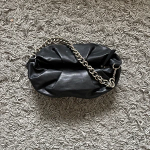 Svart axelväska med kedja - Snygg svart axelväska i skinn med en robust silverfärgad kedja som axelrem. Väskan har en dragkedja upptill och ett stilrent mönstrat innerfoder. 