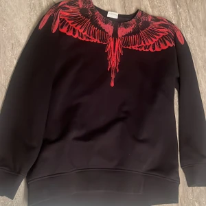 Svart tröja med röd vingdesign från Marcelo Burlon - Säljer en svart tröja från Marcelo Burlon med en cool röd vingdesign runt halsen. Tröjan har långa ärmar och är tillverkad i 100% bomull. Perfekt för dig som vill ha en unik och stilren look. Storleken är XL men den sitter som L