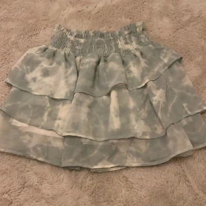 Grön och vit kjol från H&M - Säljer en söt grön och vit kjol från H&M med volanger och ett elastiskt midjeband. Kjolen har ett mönster som påminner om tie-dye och är perfekt för en avslappnad stil. Den är kort och luftig, vilket gör den idealisk för varmare dagar. Har dock en liten fläck på baksidan.