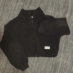 Svart teddy från Urban Classics - Mysig svart teddyjacka/tröja med dragkedja och hög krage. Perfekt för kyliga dagar. Jackan har en liten etikett på framsidan och långa ärmar med ribbade muddar.