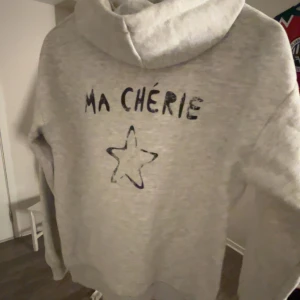 Grå hoodie med text och stjärna - Säljer en grå hoodie med texten 'MA CHÉRIE' och en stjärna tryckt på både fram- och baksidan. Tröjan har en klassisk huva och långa ärmar, perfekt för en avslappnad stil