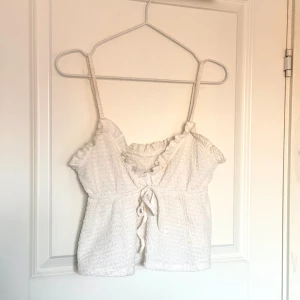 Vit ribbad topp  - Säljer en söt vit ribbad topp med smala axelband och knytning framtill. Toppen har en volangdetalj