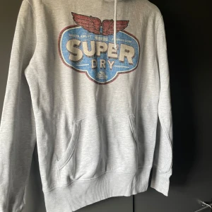 Grå hoodie från Superdry - Säljer en grå hoodie från Superdry med ett stort tryck i blått och rött på framsidan. Tröjan har en klassisk känguruficka och ribbade muddar. Perfekt för en avslappnad stil.