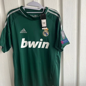 Grön Real Madrid fotbollströja från Adidas - Säljer en grön Real Madrid fotbollströja från Adidas med korta ärmar. Tröjan har vit text och klubbmärke på framsidan samt spelarnamn och nummer 7 på baksidan. Champions League-märke på ärmen. Perfekt för fans! 💚