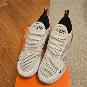 Vita Nike Air Max 270 - Snygga vita Nike Air Max 270 sneakers med svart häl och tydlig logga på plösen. Skorna har en modern design med ventilerande meshmaterial och en bekväm passform. Perfekta för både träning och vardag.för mer bilder så säg till