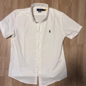 Vit kortärmad skjorta från Ralph Lauren - Säljer en stilren vit kortärmad skjorta från Ralph Lauren med klassisk krage och knappar framtill. Skjortan har en broderad logotyp på bröstet. Perfekt för en avslappnad men ändå elegant look.