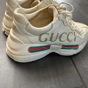 Vita sneakers från Gucci - Säljer ett par vita sneakers från Gucci med märkets logotyp i guld på sidan. Skorna har en tjock sula och klassiska snören. De har också de ikoniska gröna och röda ränderna på sidan.