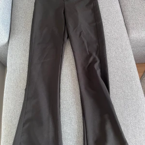 Svarta byxor från Gina Tricot - Snygga svarta byxor från Gina Tricot i storlek XS. Byxorna har en bootcut-stil med låg midja och stängs med två knappar och dragkedja. De har också praktiska bakfickor med knappar. Perfekta för en stilren look!