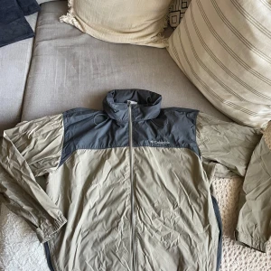 Beige och grå vindjacka från Columbia - Säljer en beige och grå vindjacka från Columbia. Jackan har en dragkedja framtill och en hög krage. Perfekt för blåsiga dagar och har en sportig look. Den är lätt och smidig att bära med sig.