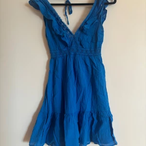 Blå klänning med spetsdetaljer - Snygg blå klänning från Calzedonia med v-ringning och spetsdetaljer. Klänningen har en knytning i ryggen och volangdetaljer vid fållen. Perfekt för sommarens alla tillfällen! Aldrig använd