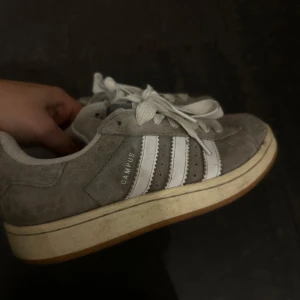 Grå Adidas Campus sneakers - Säljer ett par klassiska grå Adidas Campus sneakers med vita detaljer och snörning. Skorna har en stilren design med de ikoniska tre ränderna på sidan och en bekväm sula. Skorna är fortfarande i mycket bra skick❤️Pris går att diskutera.
