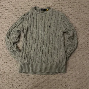 Grå kabelstickad tröja från Polo Ralph Lauren - Säljer en stilren grå kabelstickad tröja från Polo Ralph Lauren. Tröjan har en klassisk design med rund hals och det ikoniska broderade logotypen på bröstet. Perfekt för kyligare dagar och ger en tidlös look. Som är i nytt skick