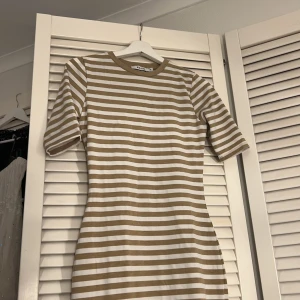 Randig klänning från NAKD - Säljer en snygg randig klänning från NAKD i beige och vit. Klänningen har korta ärmar och en normal passform. Perfekt för en avslappnad stil.