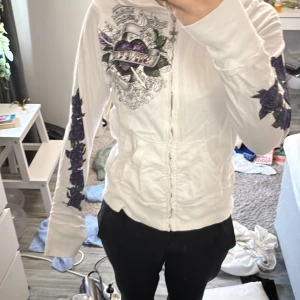 Vit hoodie med lila detaljer - Säljer en vit hoodie med dragkedja och unika lila och svarta tryck av rosor och ett hjärta. Hoodien har långa ärmar och en cool design med glitterdetaljer. Perfekt för dig som vill sticka ut!