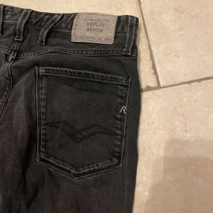 Replay Anbass W31 L34 - Snygga svarta jeans från Replay med klassisk femficksdesign och knappgylf. Perfekta för en stilren look. Märkesdetaljer på bakfickan och vid myntfickan. W31 L34