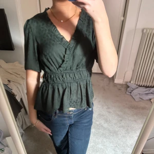 Grön omlottblus från River Island - Säljer en snygg grön omlottblus från River Island med korta ärmar och ett diskret mönster. Blusen har en smickrande v-ringning och elastisk midja för en fin passform. Perfekt för både vardag och speciella tillfällen.