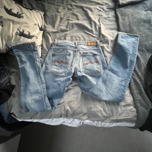Blå jeans från Nudie Jeans - Säljer ett par snygga blå jeans från Nudie Jeans med klassisk femficksdesign och orange sömmar. Jeansen har en broderad detalj på bakfickorna och är tillverkade i ekologisk bomull. Perfekta för dig som gillar en stilren look!
