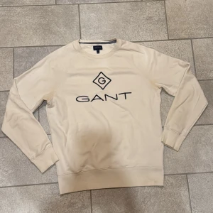 Vit sweatshirt från GANT - Säljer en stilren vit sweatshirt från GANT med deras ikoniska logga tryckt på framsidan. Tröjan har långa ärmar och en klassisk rund halsringning. Perfekt för en avslappnad och trendig look.