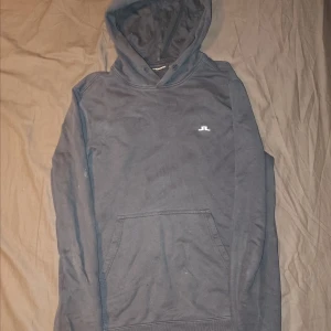 Mörkblå hoodie från J.Lindeberg - Säljer en stilren mörkblå hoodie från J.Lindeberg med en klassisk känguruficka och justerbar huva. Den har en liten logotyp på bröstet. Perfekt för en avslappnad look. Den är väl använd och har sina defekter som visas i bilderna