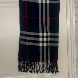 Rutig halsduk med fransar - Snygg rutig halsduk i mörkblått med vita, svarta och röda ränder. Den har fransar i ändarna som ger en extra touch. Perfekt för att hålla sig varm och stilren under kyliga dagar.