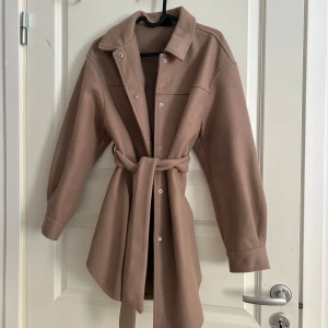 Beige kappa från Vero Moda - Snygg beige kappa från Vero Moda med knappar och bälte i midjan. Perfekt för höst och vår. Den har långa ärmar och en klassisk krage.