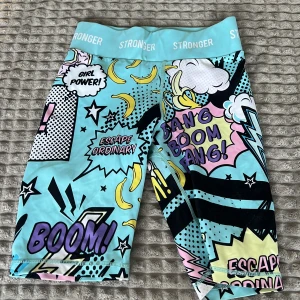 Färgglada träningsshorts från Stronger - Snygga och färgglada träningsshorts från Stronger med serietidningsinspirerat mönster. De har en hög midja och är perfekta för att sticka ut på gymmet. Shortsen har text och grafik i olika färger som blå, gul och rosa.