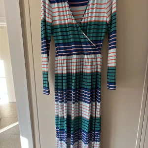 Randig omlottklänning från Stine goya  - Snygg långärmad omlottklänning med randigt mönster i grönt, blått, vitt och orange. Perfekt för en färgglad look. I viskos!! 