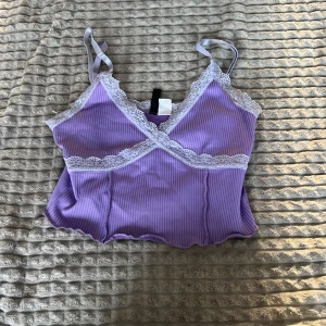 Lila ribbad linnetopp med spetsdetaljer - Säljer en söt lila ribbad linnetopp med vita spetsdetaljer. Toppen har smala axelband och en omlott-design framtill. Perfekt för varma dagar eller som ett lager under en tröja.