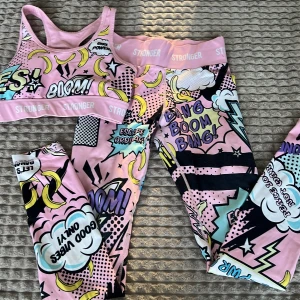 Färgglatt träningsset från Stronger - Säljer ett färgglatt träningsset från Stronger med ett coolt serietidningsmönster. Setet består av en rosa sport-bh och matchande tights med text och bananmönster. Perfekt för dig som vill sticka ut på gymmet! 💪