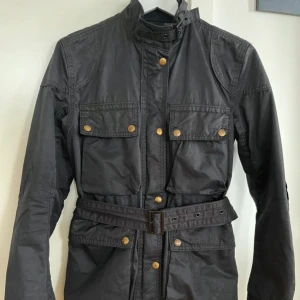 Ralph Lauren jacka - Snygg svart jacka från Denim & Supply Ralph Lauren med knappar och bälte i midjan. Jackan har långa ärmar och flera fickor med knappstängning. Perfekt för en stilren look. Storlek XS.