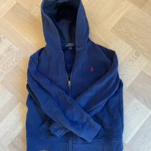 Mörkblå hoodie från Ralph Lauren - Säljer en snygg mörkblå hoodie från Ralph Lauren med dragkedja och huva. Den har en liten röd logga på bröstet och praktiska fickor framtill. Perfekt för en avslappnad stil. Tecken på användning men fortfarande i fint skick. Barnstorlek L som motsvarar ungefär xs. Passar dig mellan 160-170! Vid frågor är de som vanligt bara att kontakta mig👍 