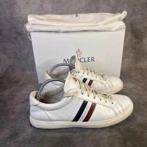 Moncler monaco sko - Snygga vita sneakers från Moncler med klassiska ränder i rött och blått på sidan. Skorna har snörning och är tillverkade i skinn, vilket ger en lyxig känsla. Perfekta för en stilren look. Storleken säger 43 men passar 42.