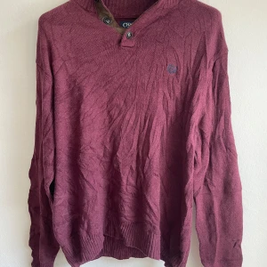 Vinröd tröja från Chaps - Säljer en vinröd tröja från Chaps med knappar vid halsen och bruna armbågslappar. Tröjan har långa ärmar och ribbstickade muddar vid ärmslut och nederkant. Perfekt för kyligare dagar!