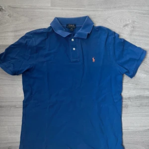 Blå pikétröja från Ralph Lauren - Säljer en klassisk blå pikétröja från Ralph Lauren med korta ärmar och en liten broderad logga på bröstet. Perfekt för en stilren och avslappnad look. Tröjan har en krage och knappar framtill. Tröjan passar M/S va inte rädda för att skriva över minsta tanken 
