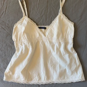 Vit linnetopp från Brandy Melville - Säljer en söt vit linnetopp från Brandy Melville med spetsdetaljer vid halsringning och nederkant. Toppen har smala axelband och en v-ringad design. Perfekt för varma dagar!