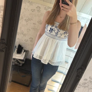 Vit ärmlös blus med blå broderier - Säljer en söt vit blus med blå broderier och smala axelband. Blusen har en luftig passform och ett fint mönster upptill. Perfekt för varma dagar!